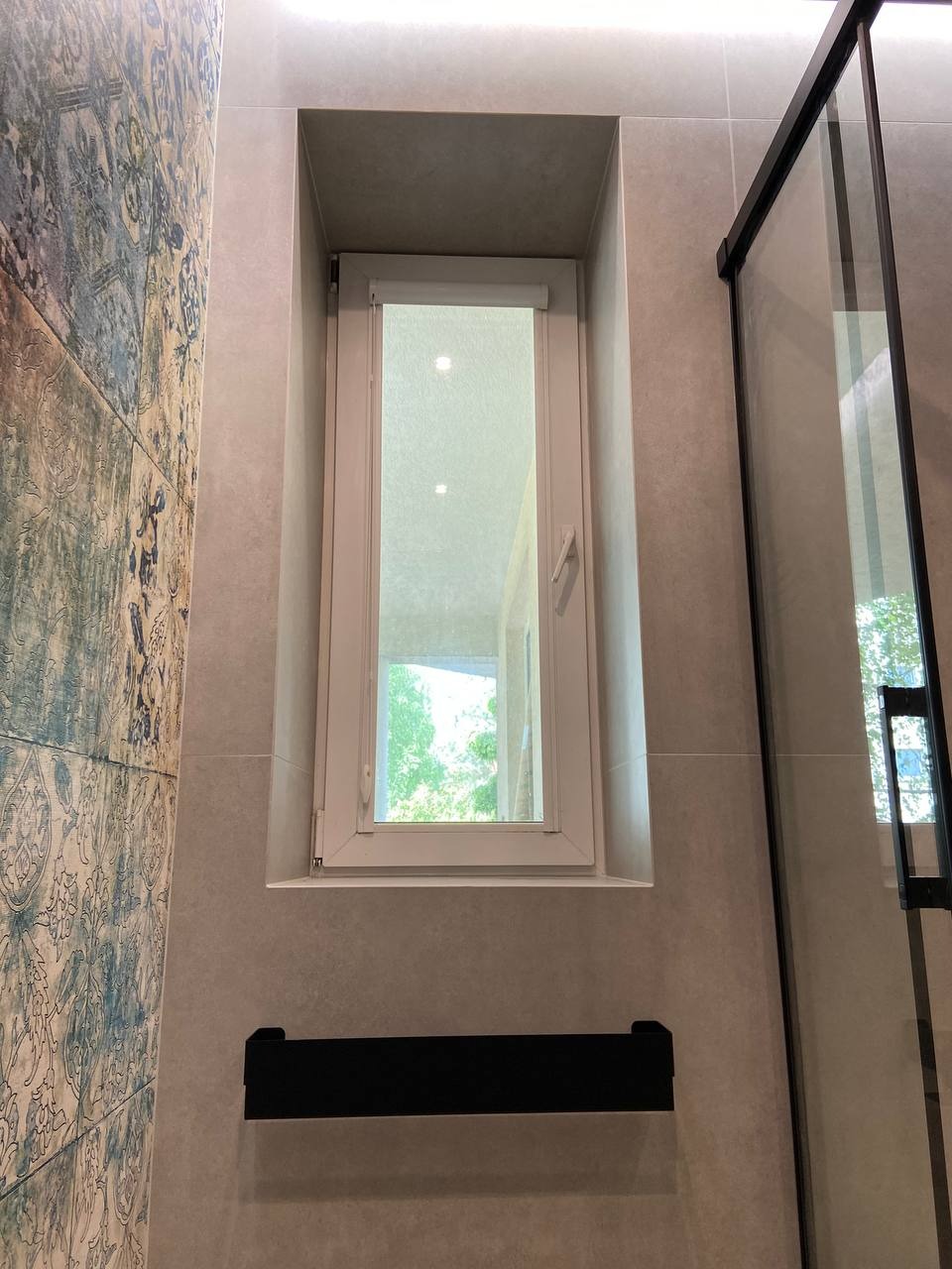 Wąskie okno z roletą wnękowe w łazience, obok kabina prysznicowa z czarnymi ramami, ściana w szaro-niebieskie wzorzyste płytki.