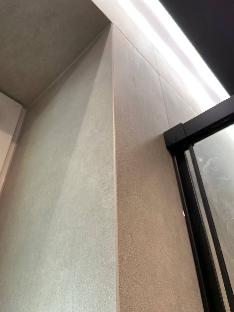 Fragment narożnika łazienki z jasnymi, matowymi płytkami ceramicznymi, widoczna czarna rama kabiny prysznicowej i oświetlenie podsufitowe LED.