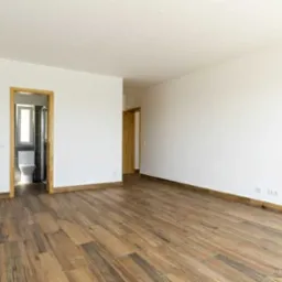 Pusty pokój z drewnianą podłogą ułożoną w jodełkę, białe ściany i widok na łazienkę z toaletą przez otwarte drzwi.