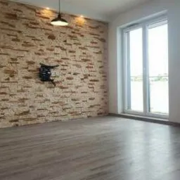 Wykończone wnętrze pokoju z kamienną ścianą dekoracyjną, panelami podłogowymi w odcieniach szarości oraz uchwytem na telewizor zamontowanym na ścianie.