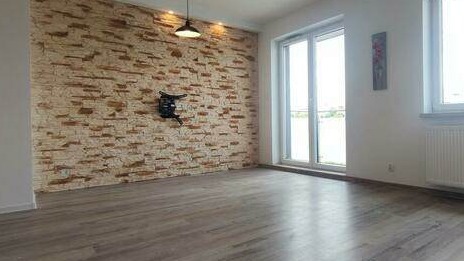 Wykończone wnętrze pokoju z kamienną ścianą dekoracyjną, panelami podłogowymi w odcieniach szarości oraz uchwytem na telewizor zamontowanym na ścianie.