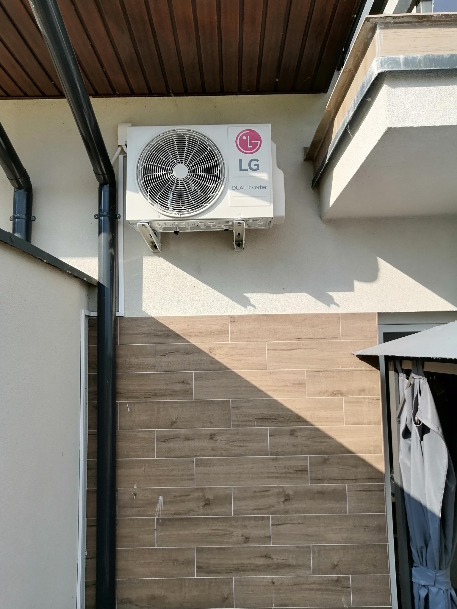 Zewnętrzna jednostka klimatyzacji LG Dual Inverter zamontowana na elewacji z drewnopodobnych płytek, widoczny fragment rynny i zadaszenia.
