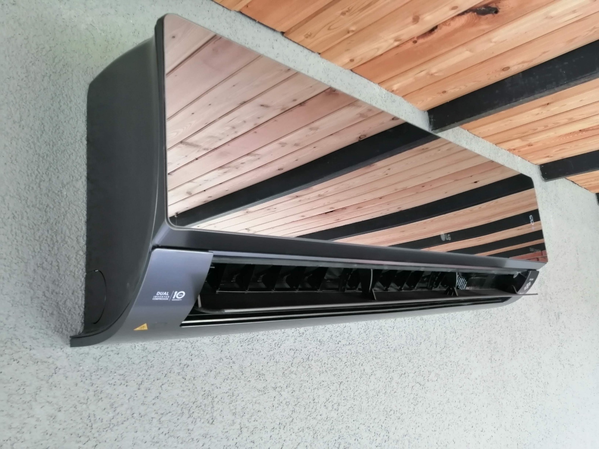 Szary klimatyzator LG zamontowany na teksturowanej ścianie, z widocznym drewnianym sufitem odbijającym się w jego błyszczącej obudowie.