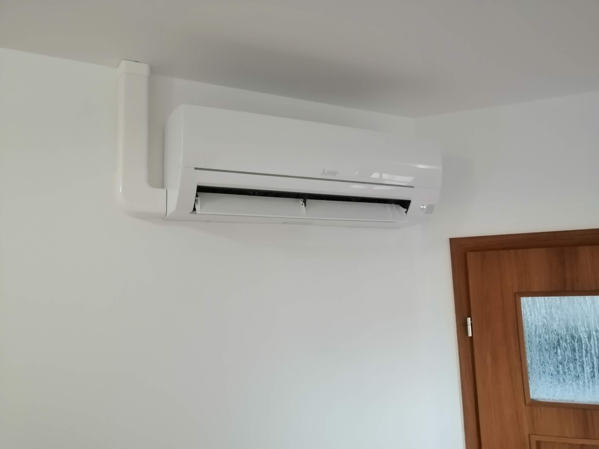 Biały klimatyzator Mitsubishi zamontowany na białej ścianie w pokoju, widoczna brązowa drewniana rama drzwi z szybą w tle.