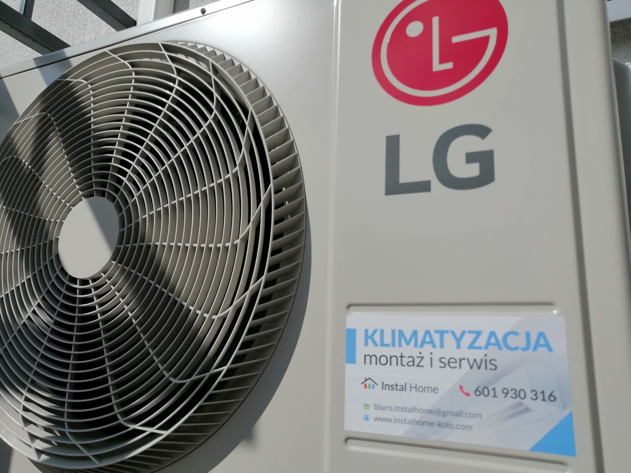 Zewnętrzna jednostka klimatyzacji LG z widocznym wentylatorem i naklejką firmy oferującej montaż i serwis, zamontowana na elewacji budynku.