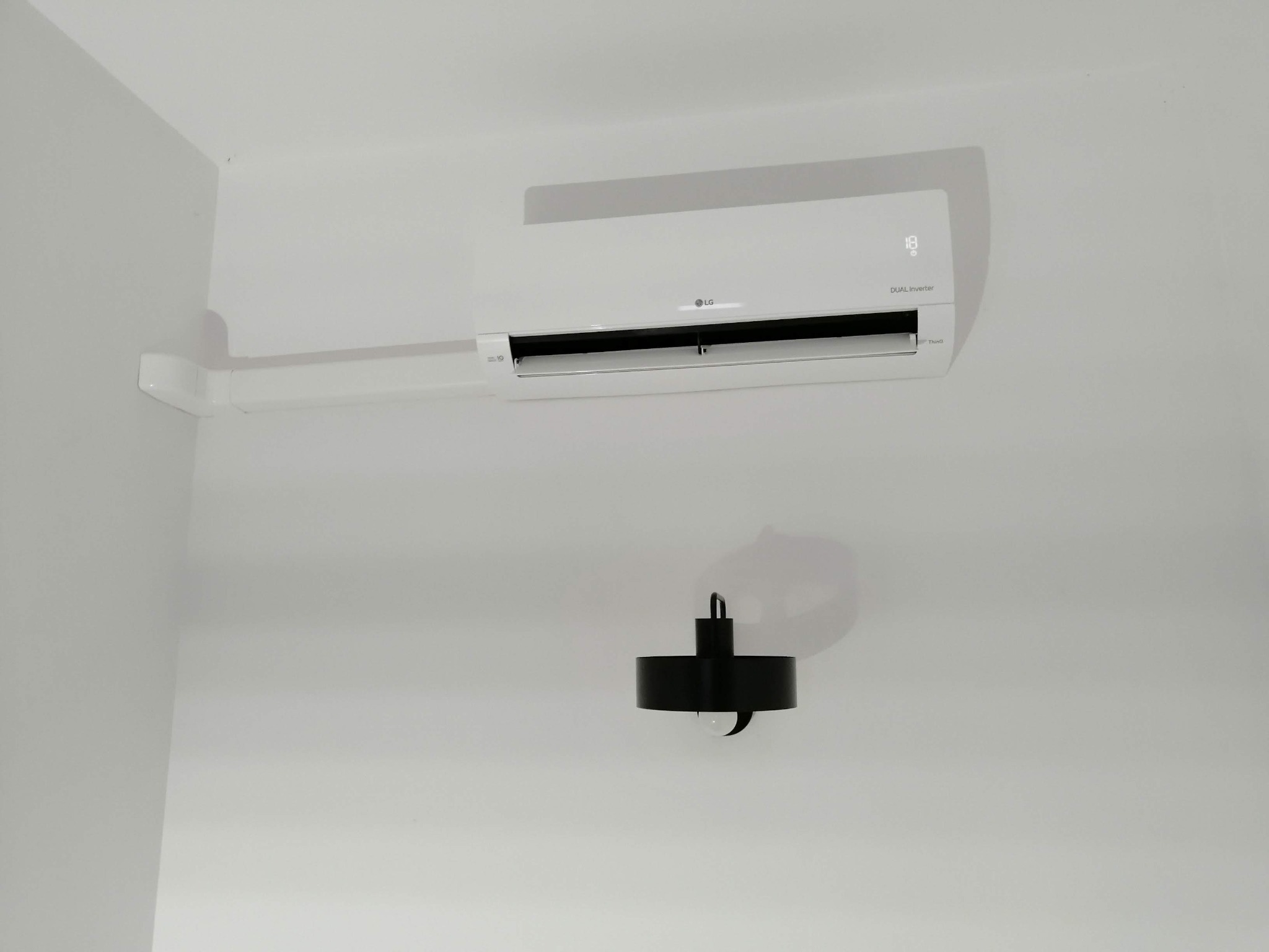 Biały klimatyzator LG Dual Inverter zamontowany na białej ścianie, z widocznym wyświetlaczem temperatury i czarną lampą ścienną poniżej.