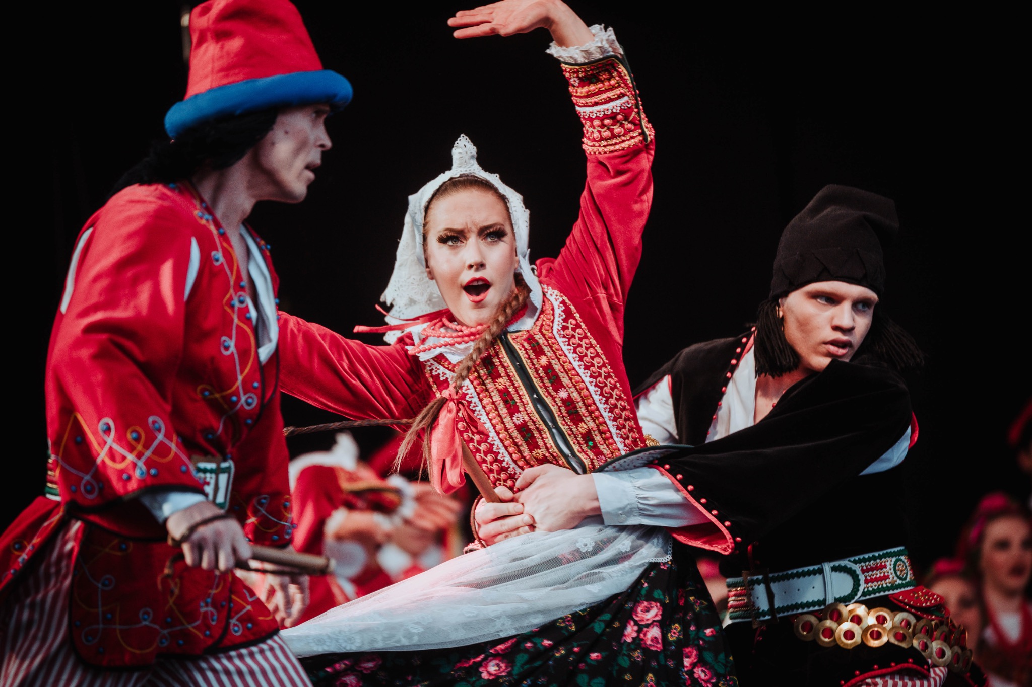 Zespół ludowy w tradycyjnych, kolorowych strojach podczas dynamicznego występu na scenie, oświetlony reflektorami.