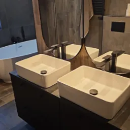 Nowoczesna łazienka z owalnym lustrem, dwiema białymi umywalkami na czarnej szafce i wolnostojącą wanną przy oknie. Szare płytki imitujące drewno na ścianach i podłodze.