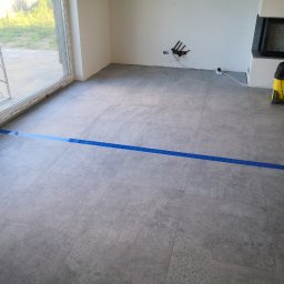 Artur Lewiński - Szare płytki podłogowe w nowym wnętrzu, zabezpieczone niebieską taśmą malarską przy oknie. Widoczne przyłącza na ścianie i odkurzacz przy kominku.