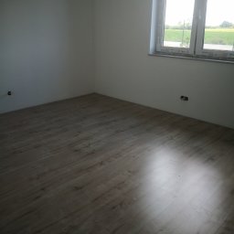 Artur Lewiński - Jasne panele podłogowe w pustym pokoju z oknem, widok na zewnątrz. Świeżo wykończone wnętrze z gładkimi, białymi ścianami i widocznymi gniazdkami elektrycznymi.