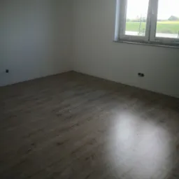 Jasne panele podłogowe w pustym pokoju z oknem, widok na zewnątrz. Świeżo wykończone wnętrze z gładkimi, białymi ścianami i widocznymi gniazdkami elektrycznymi.