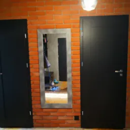 Ceglana ściana z czarnymi drzwiami po bokach i lustrem pośrodku, które odbija wnętrze pokoju. Widoczna żarówka i fragment podłogi.