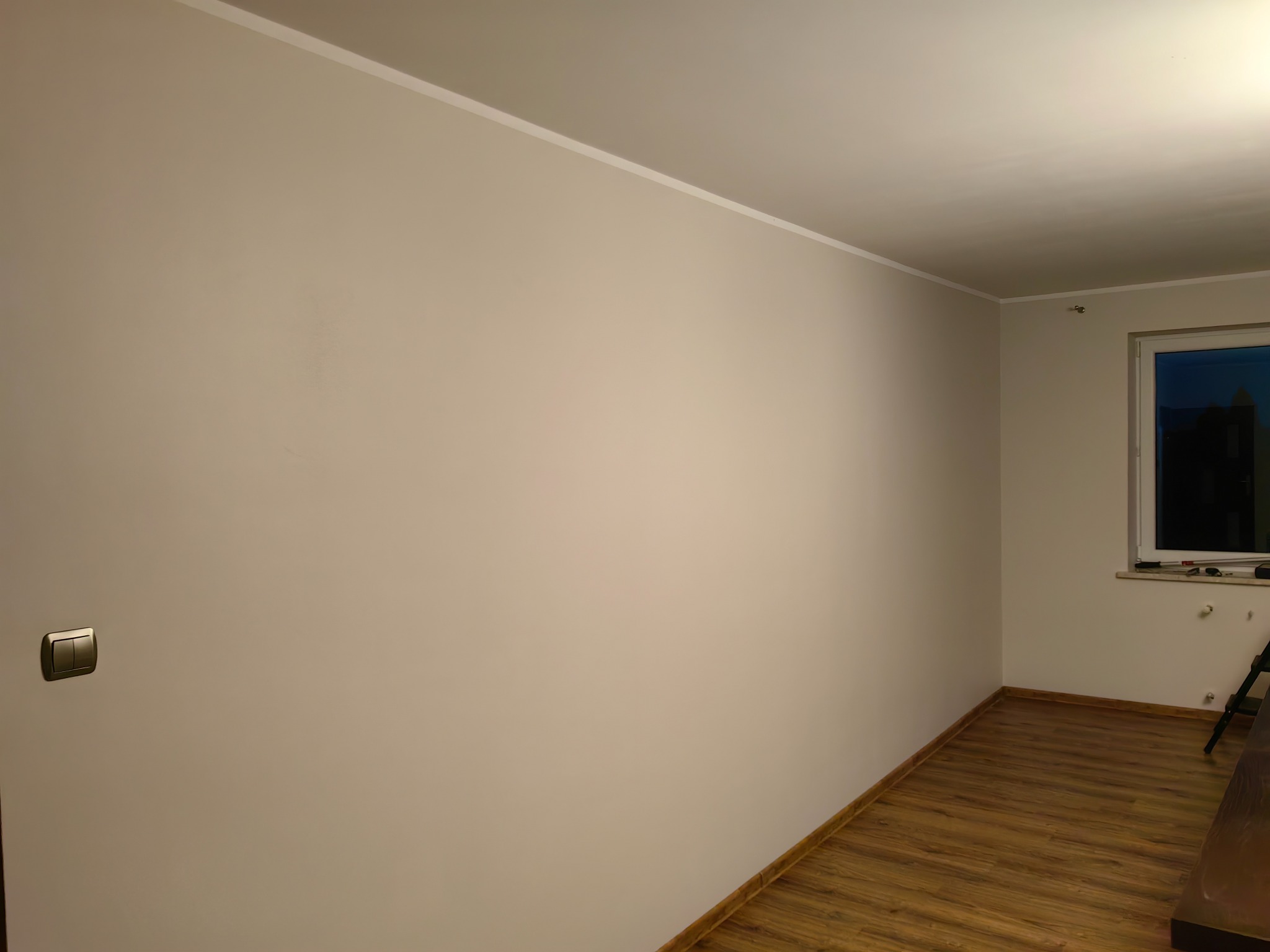 Jasny pokój po remoncie: świeżo pomalowane ściany w odcieniach beżu, drewniana podłoga, biała listwa sufitowa, okno z widokiem na zewnątrz i włącznik światła.