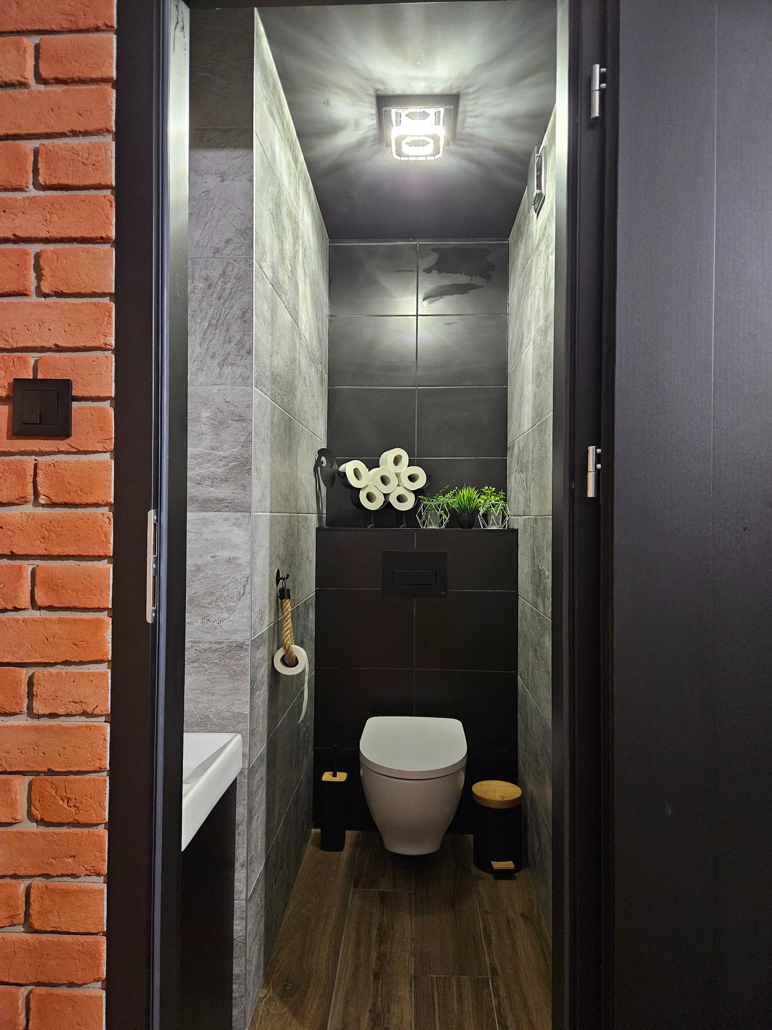 Stylowo wykończona, wąska toaleta w Raciążu. Ciemne płytki kontrastują z ceglaną ścianą zewnętrzną. Widoczne detale: drewniana podłoga i dekoracje.