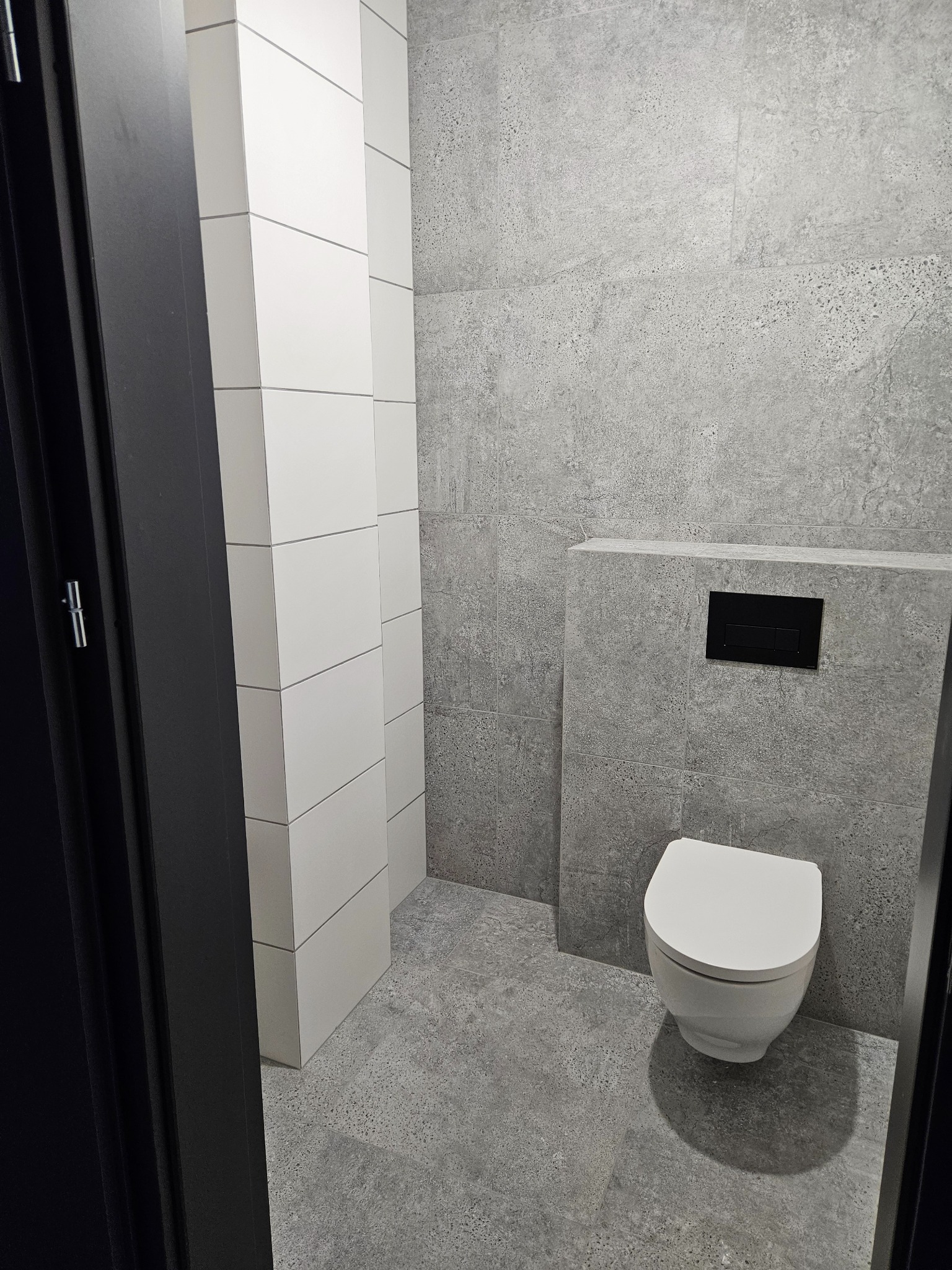 Szara łazienka po remoncie: biała wisząca toaleta, szare płytki na ścianach i podłodze, czarny panel sterujący. Widok zza czarnej futryny drzwi.