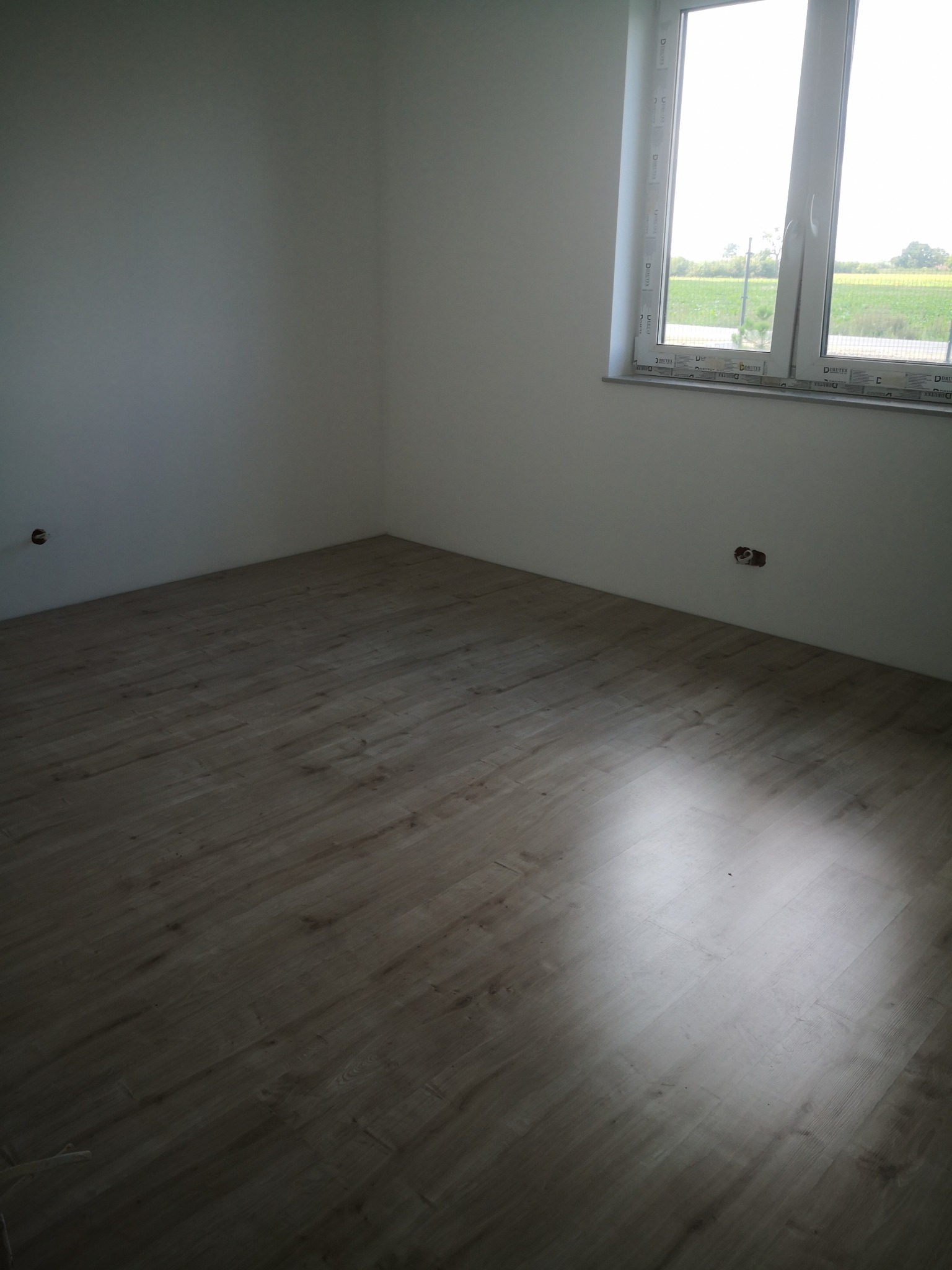 Jasne panele podłogowe w pustym pokoju z oknem, widok na zewnątrz. Świeżo wykończone wnętrze z gładkimi, białymi ścianami i widocznymi gniazdkami elektrycznymi.