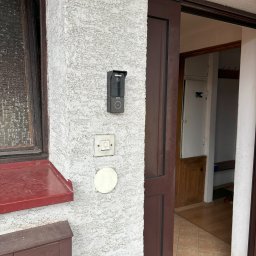 ONEXO.pl Adrian Sokół - Nowoczesny wideodomofon z kamerą zamontowany na szorstkiej ścianie obok tradycyjnego dzwonka i skrzynki na listy z symbolem poczty; widok na otwarte drzwi wejściowe.