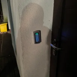 Zewnętrzny panel numeryczny domofonu z podświetlanymi przyciskami, zamontowany na szarej, chropowatej ścianie obok ciemnych drzwi wejściowych z metalową klamką.