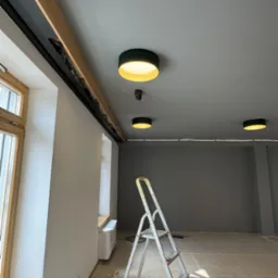 Wnętrze biura w trakcie instalacji oświetlenia: widoczne okna, sufit z zamontowanymi lampami, drabina i przewody.