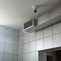 ONEXO.pl Adrian Sokół - Kamera kopułkowa zamontowana na suficie w pomieszczeniu z białą, kwadratową glazurą, widoczny przewód wentylacyjny i fragment baterii kuchennej.