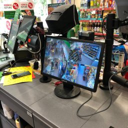 ONEXO.pl Adrian Sokół - Ekran monitora z podglądem z kamer monitoringu w sklepie spożywczym, widoczne kasy fiskalne, terminale płatnicze, telefon i notatki.