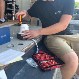 ONEXO.pl Adrian Sokół - Instalator przykręca śrubokrętem białą kamerę z logo 'kenik' do czarnej podstawy; obok otwarte pudełko z izolowanymi śrubokrętami, pudełko kamery, śruby montażowe i drabina.