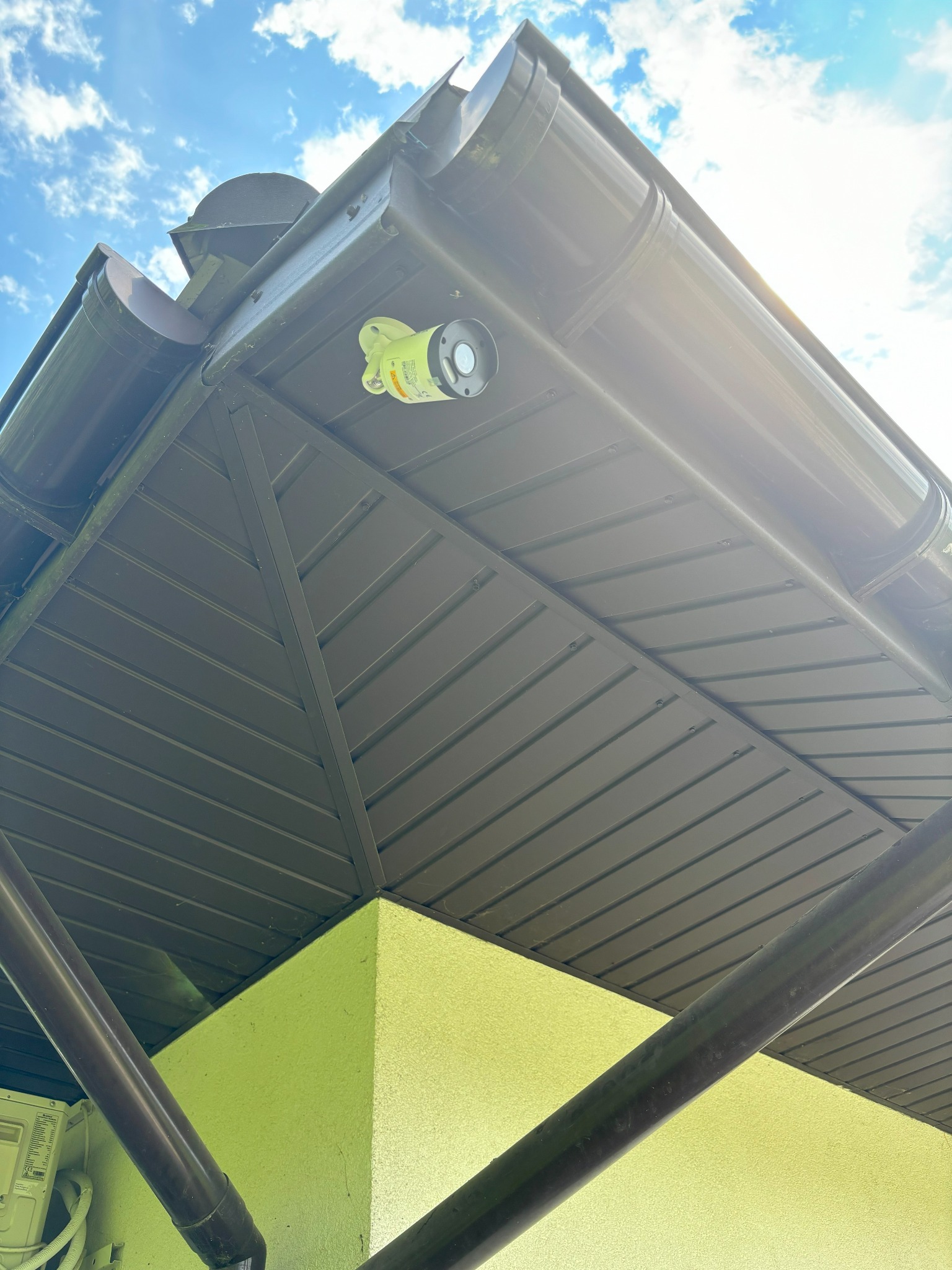 Kamera cylindryczna w obudowie w kolorze seledynowym zamontowana pod okapem dachu, widok z dołu na tle błękitnego nieba z chmurami.