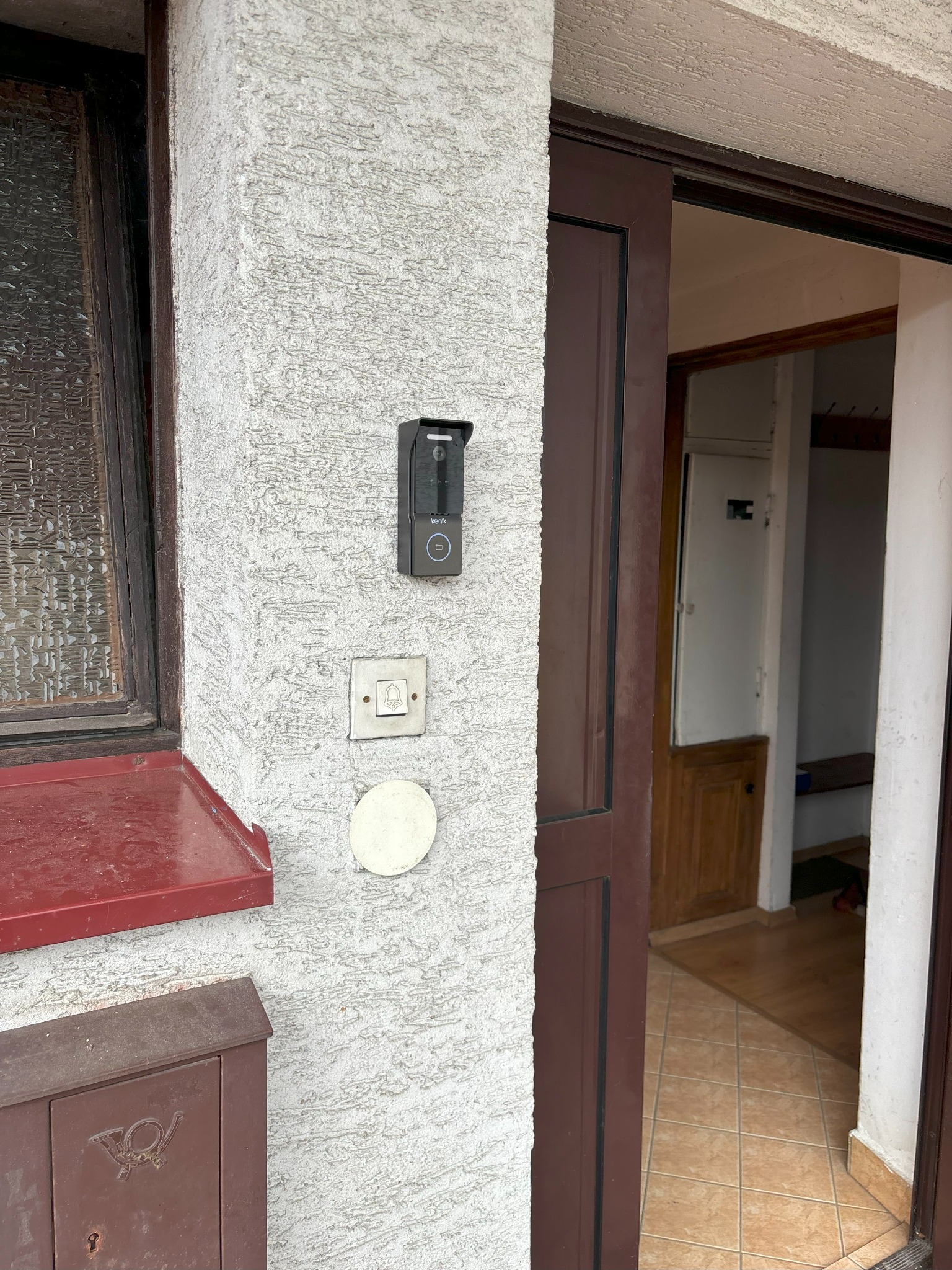 Nowoczesny wideodomofon z kamerą zamontowany na szorstkiej ścianie obok tradycyjnego dzwonka i skrzynki na listy z symbolem poczty; widok na otwarte drzwi wejściowe.