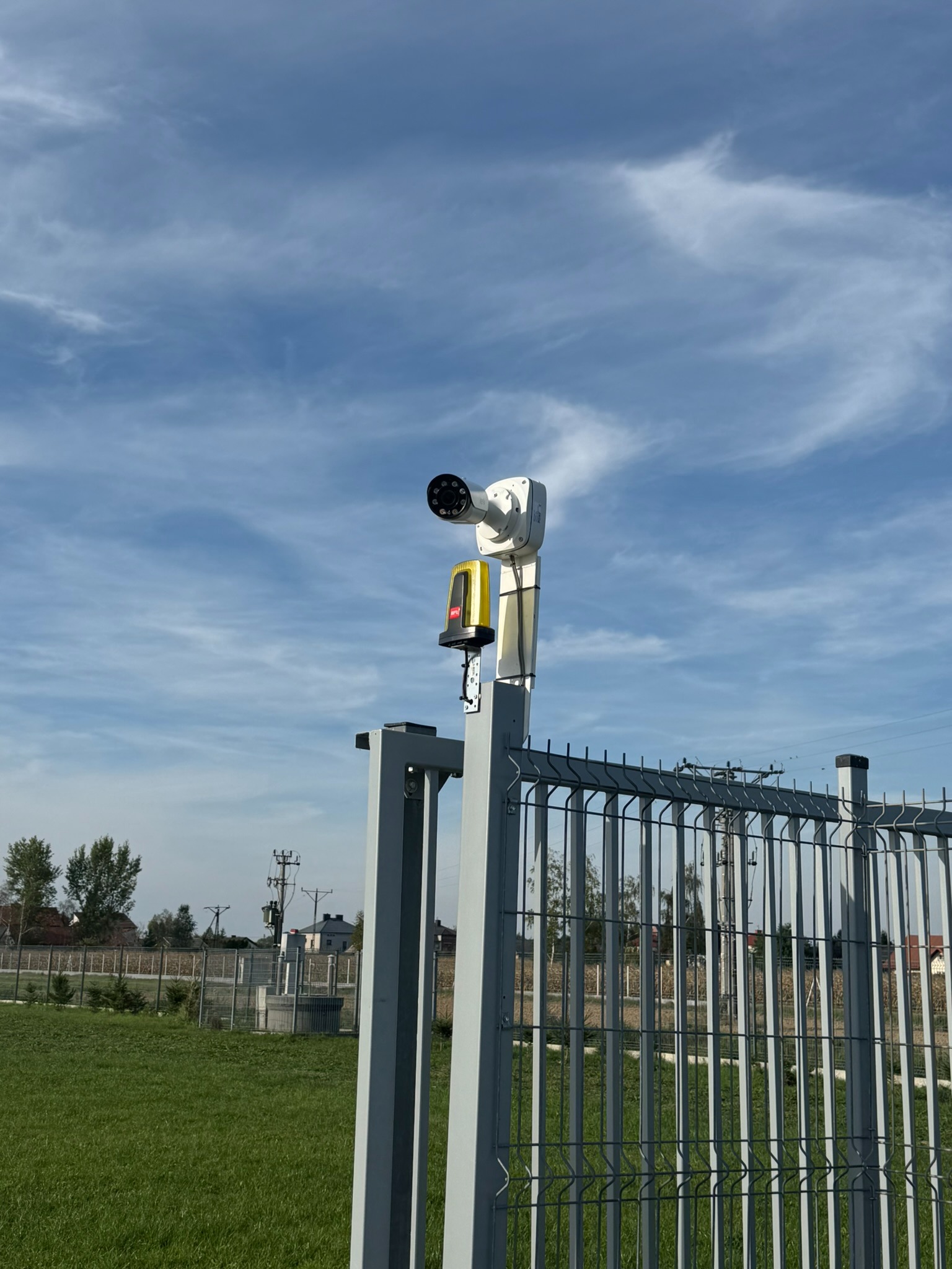 Biała kamera monitoringu z żółtym modułem oświetleniowym zamontowana na szarym słupku ogrodzenia panelowego, na tle błękitnego nieba z chmurami.