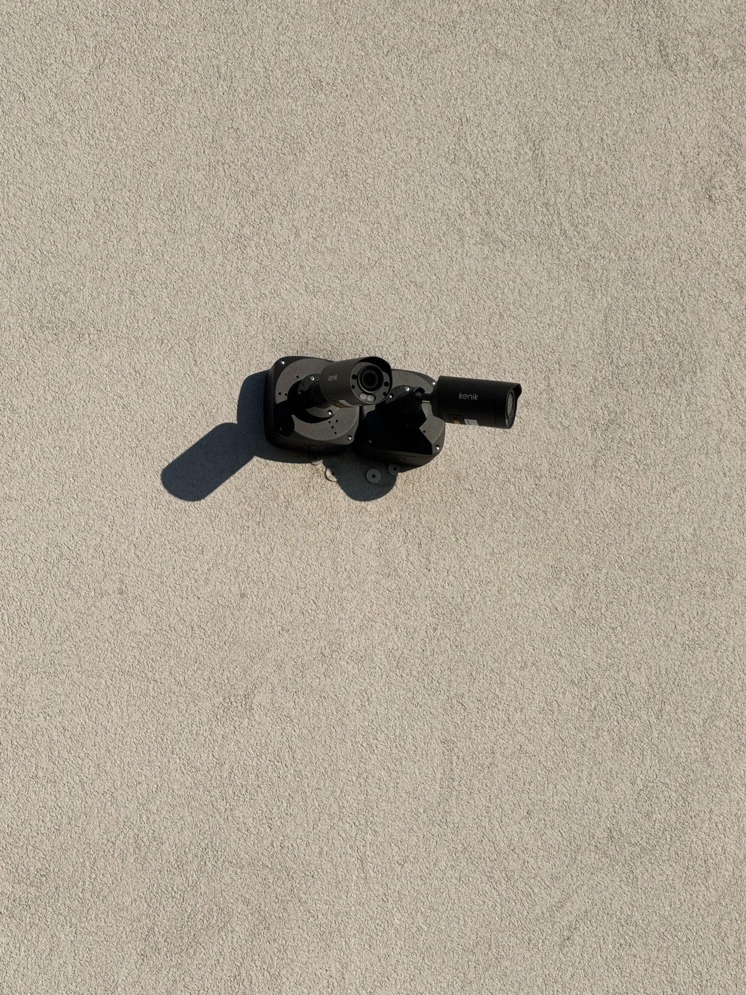 Kamera przemysłowa marki Kenik zamontowana na szarej, teksturowanej ścianie budynku, rzucająca cień po lewej stronie.