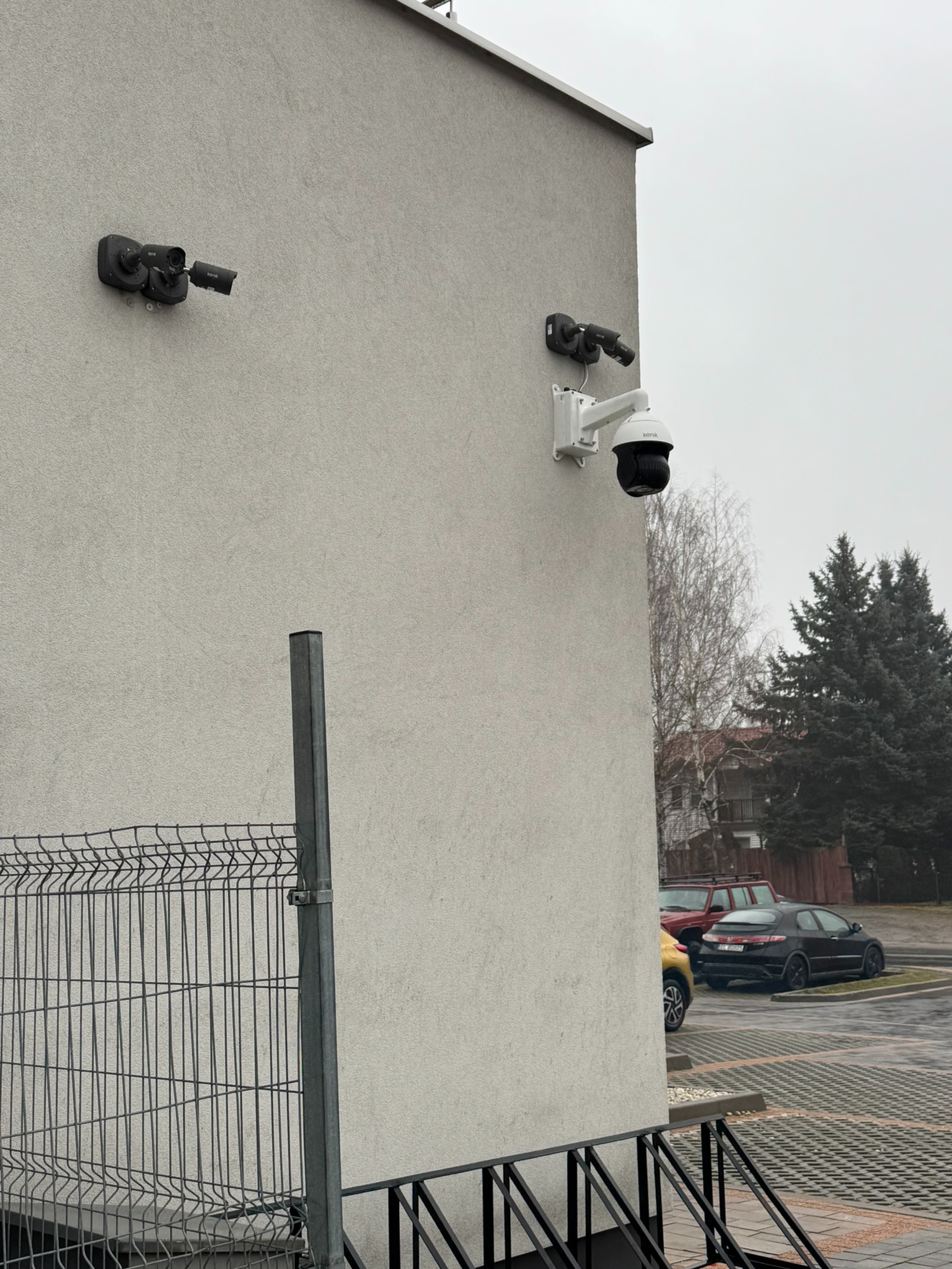Dwie kamery przemysłowe marki Kenik zamontowane na elewacji budynku, widoczne ogrodzenie panelowe i zaparkowane samochody w tle.