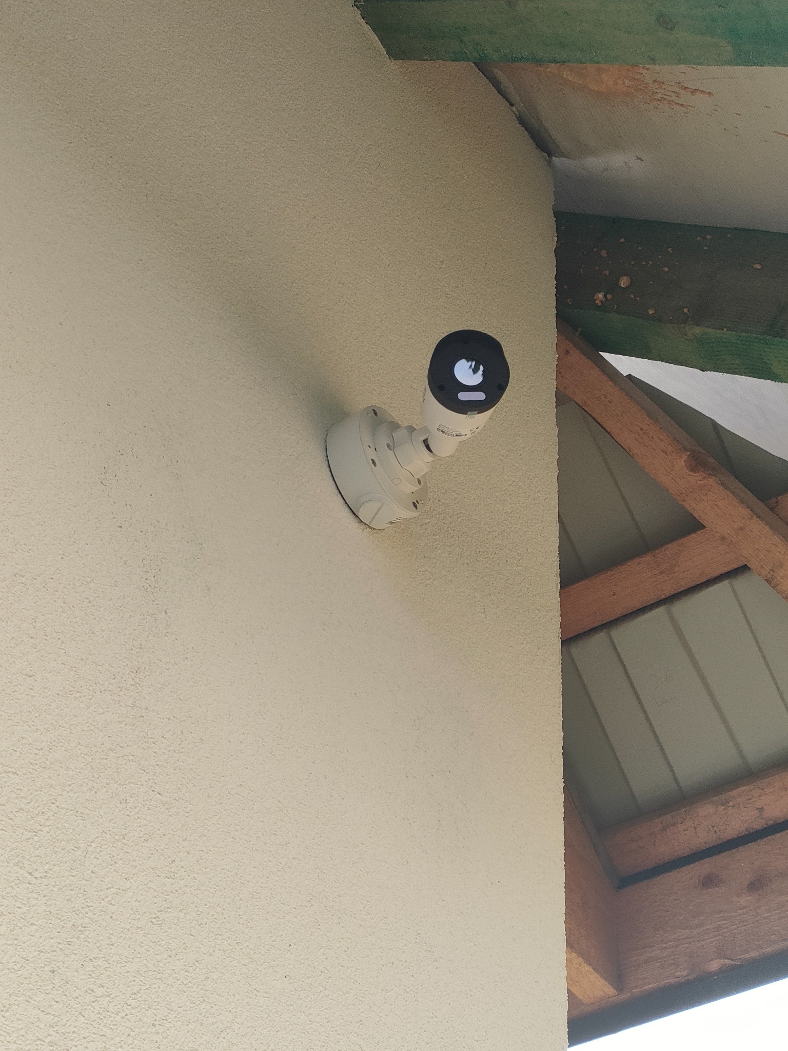 Kamera monitoringu zamontowana na jasnej, teksturowanej ścianie zewnętrznej, z widocznymi elementami drewnianej konstrukcji dachu w tle.