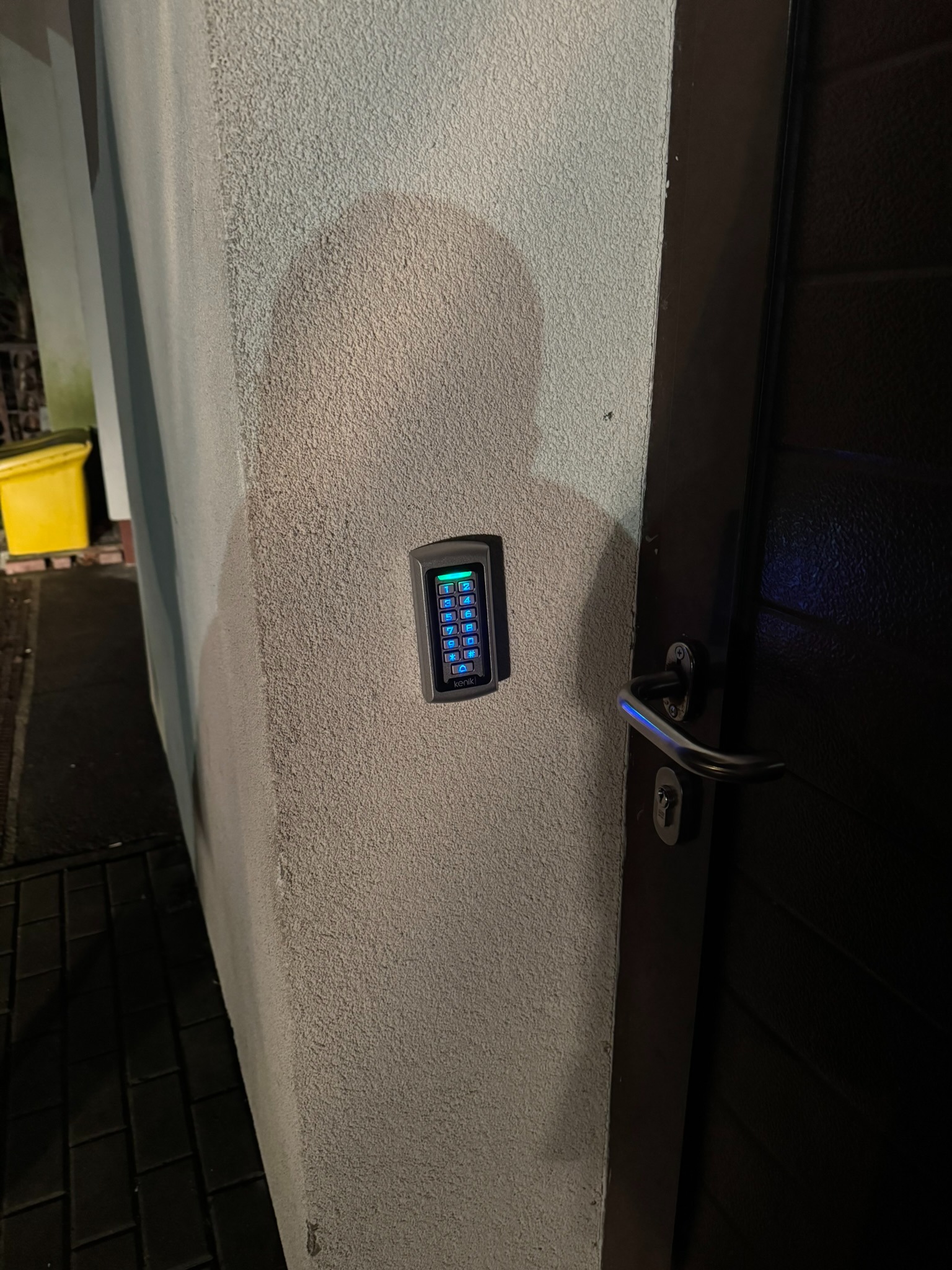 Zewnętrzny panel numeryczny domofonu z podświetlanymi przyciskami, zamontowany na szarej, chropowatej ścianie obok ciemnych drzwi wejściowych z metalową klamką.