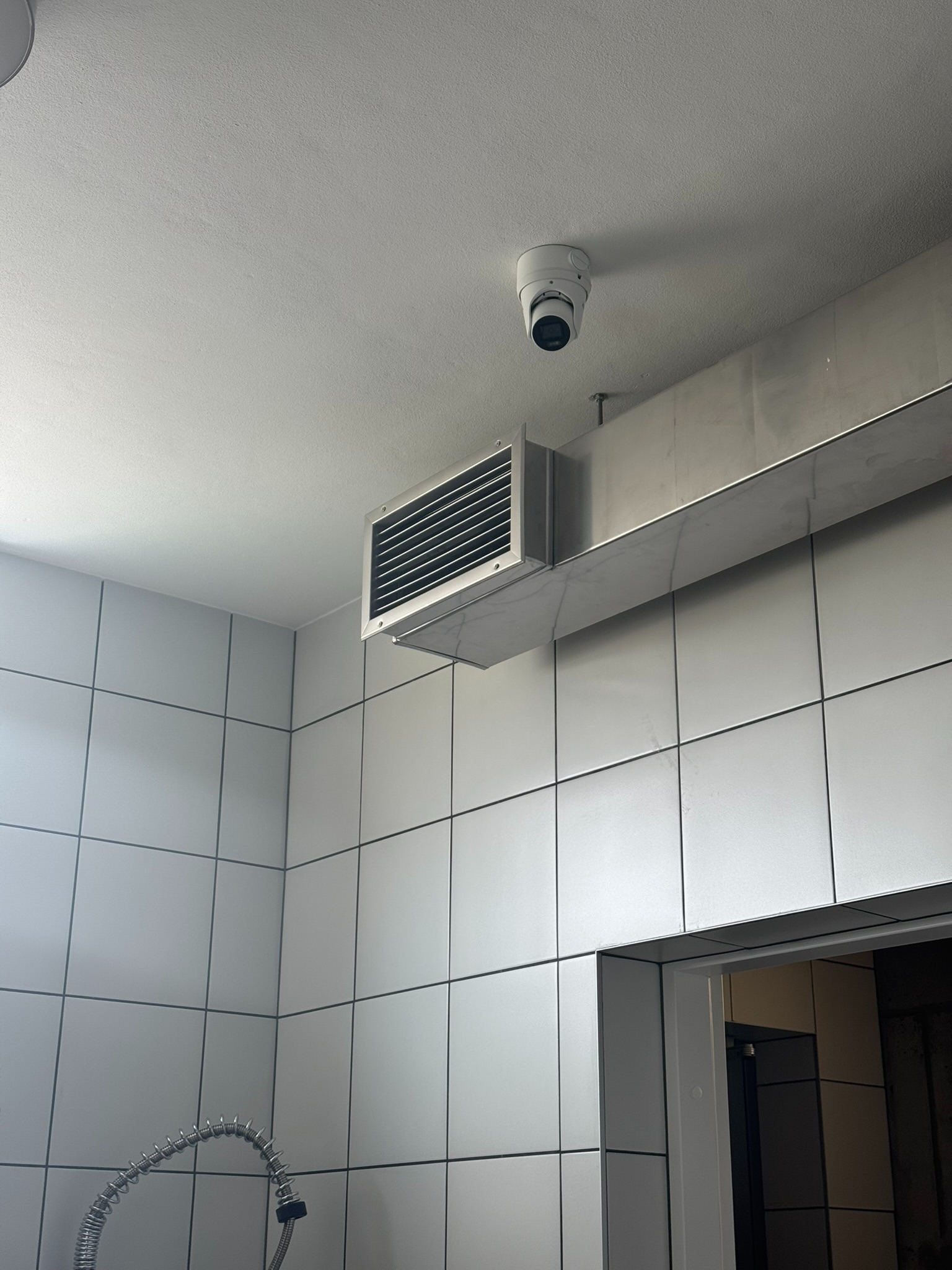 Kamera kopułkowa zamontowana na suficie w pomieszczeniu z białą, kwadratową glazurą, widoczny przewód wentylacyjny i fragment baterii kuchennej.