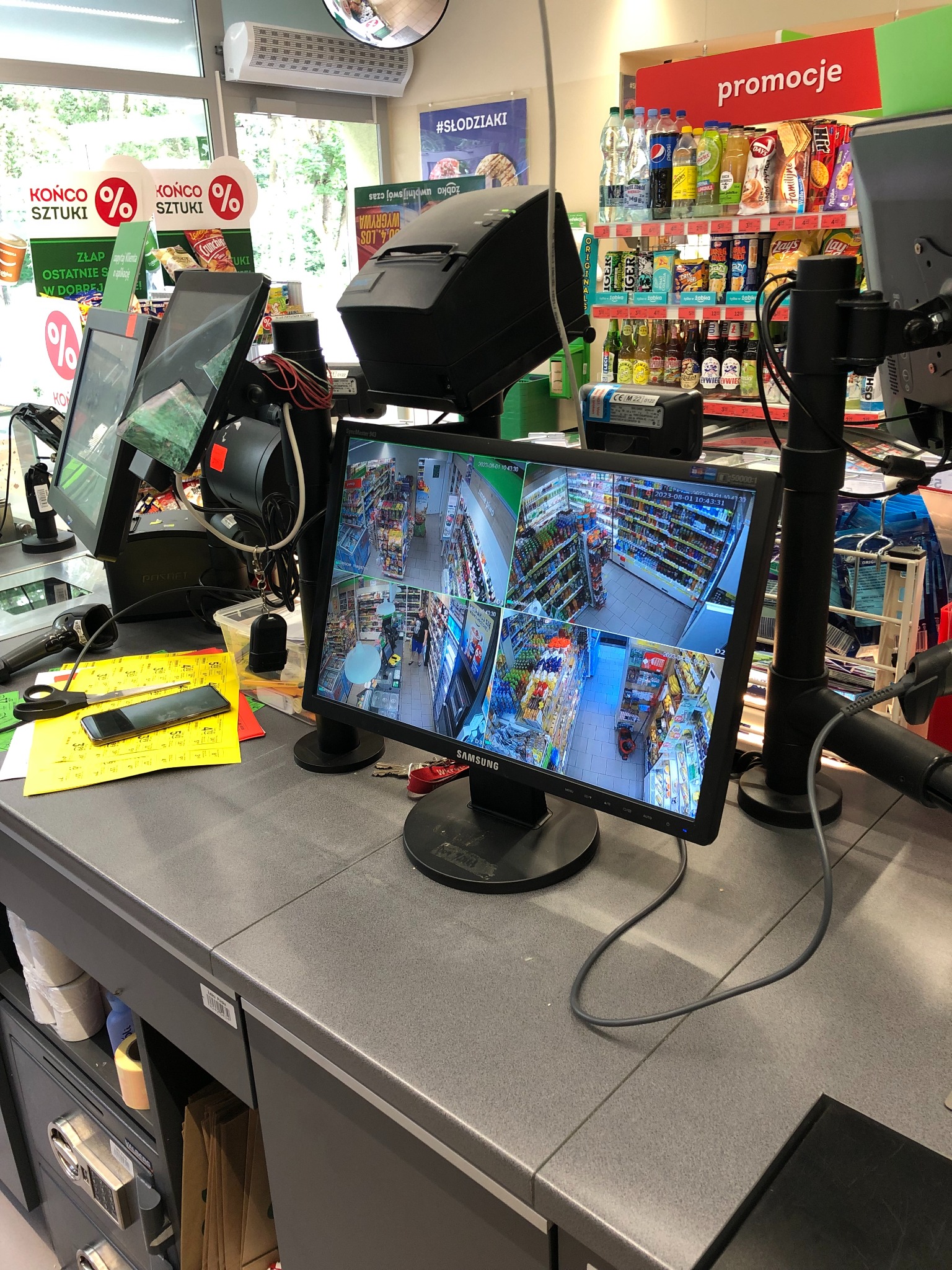 Ekran monitora z podglądem z kamer monitoringu w sklepie spożywczym, widoczne kasy fiskalne, terminale płatnicze, telefon i notatki.