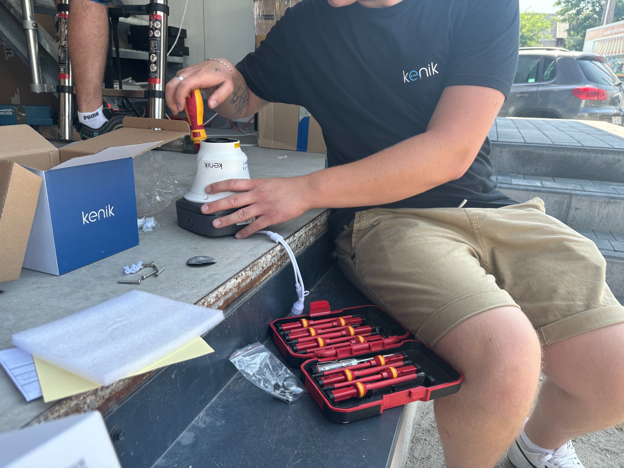 Instalator przykręca śrubokrętem białą kamerę z logo 'kenik' do czarnej podstawy; obok otwarte pudełko z izolowanymi śrubokrętami, pudełko kamery, śruby montażowe i drabina.