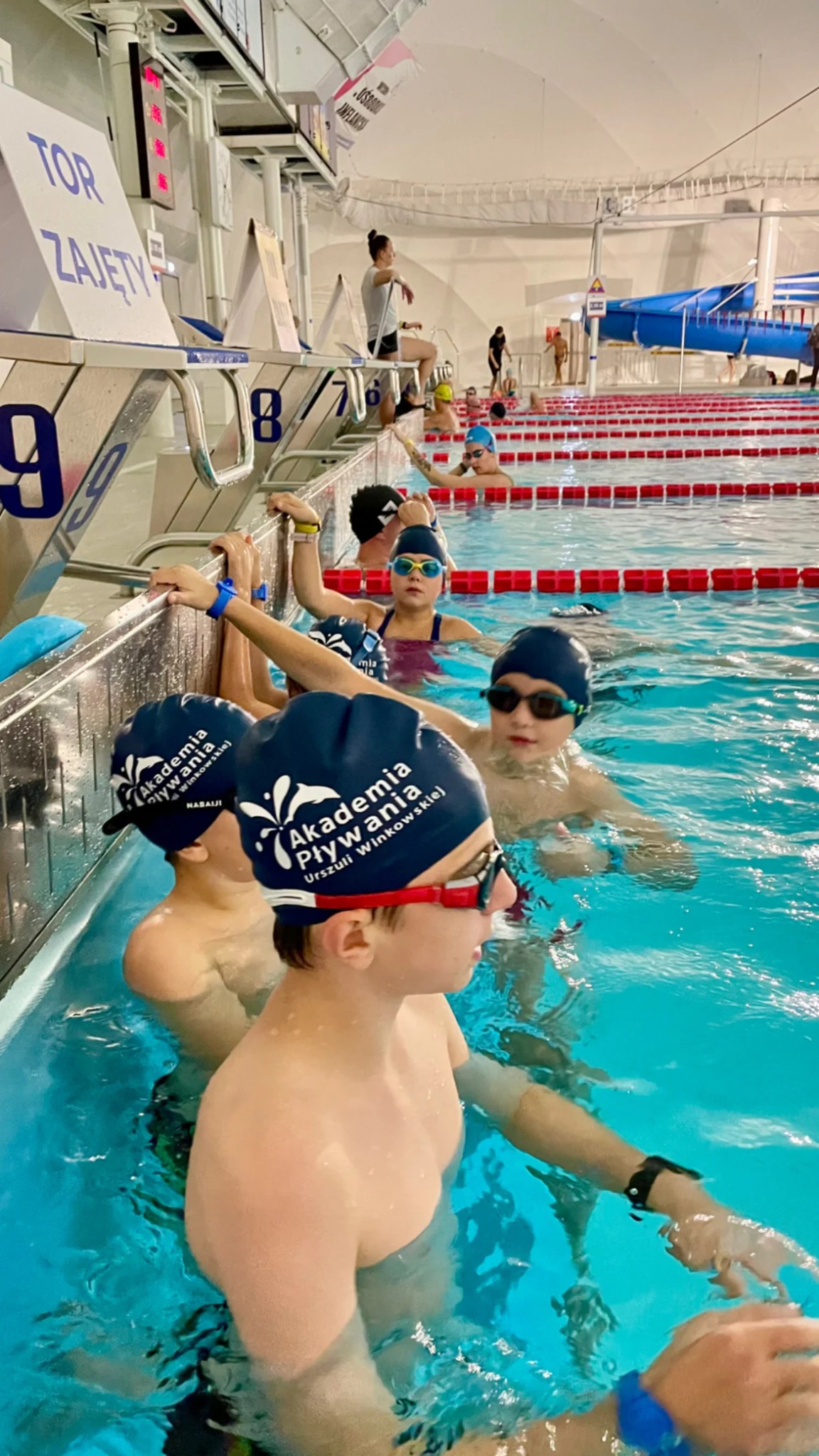 Grupa dzieci w czepkach pływackich Akademii Pływania Urszuli Winkowskiej, stojących w basenie, czekających na rozpoczęcie zajęć, z widocznymi torami i numeracją.