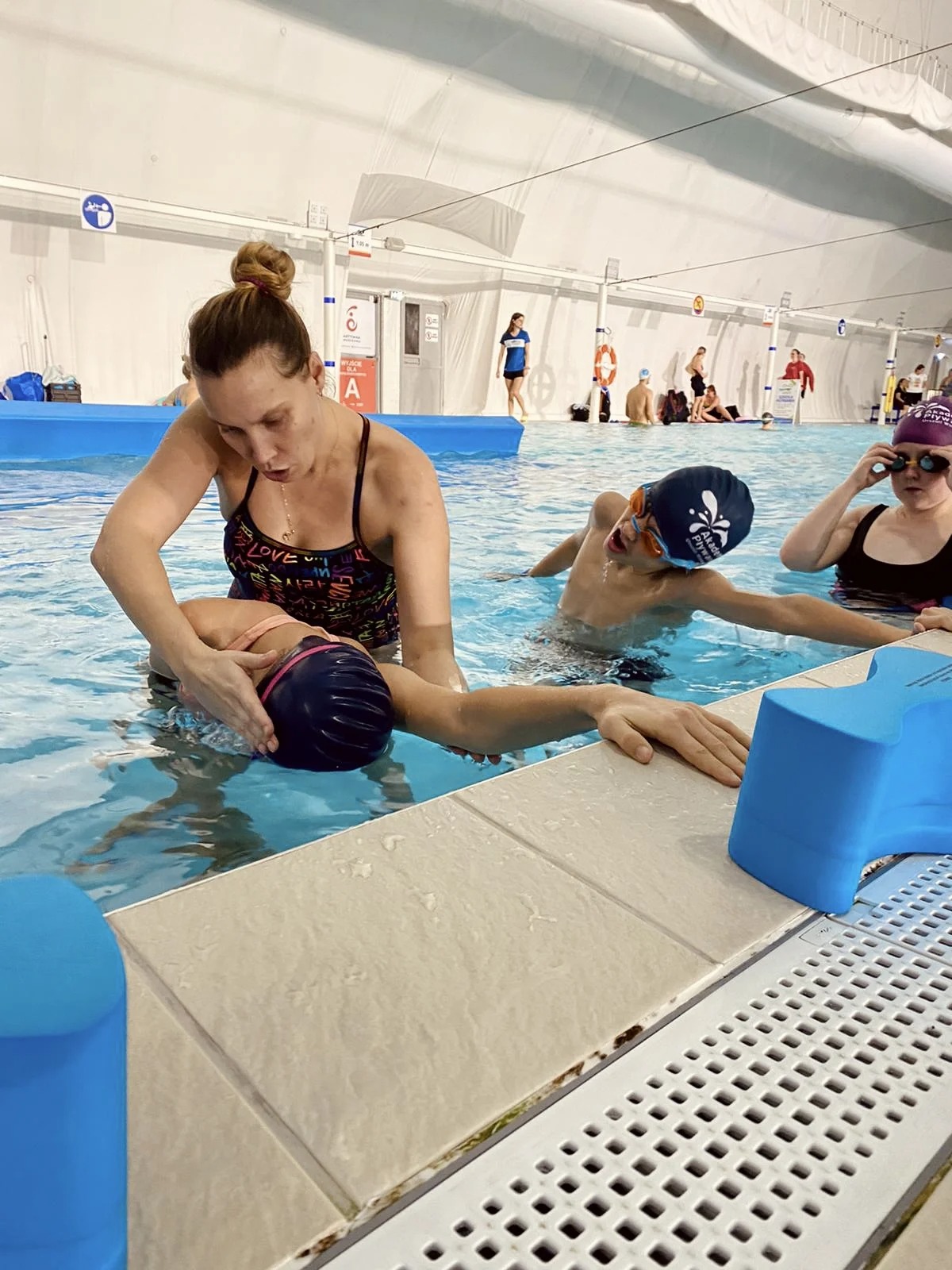 Instruktorka w stroju kąpielowym, z włosami upiętymi w kok, pomaga dziecku w czepku utrzymać się na wodzie podczas nauki pływania w basenie krytym. W tle widoczni inni uczestnicy zajęć.