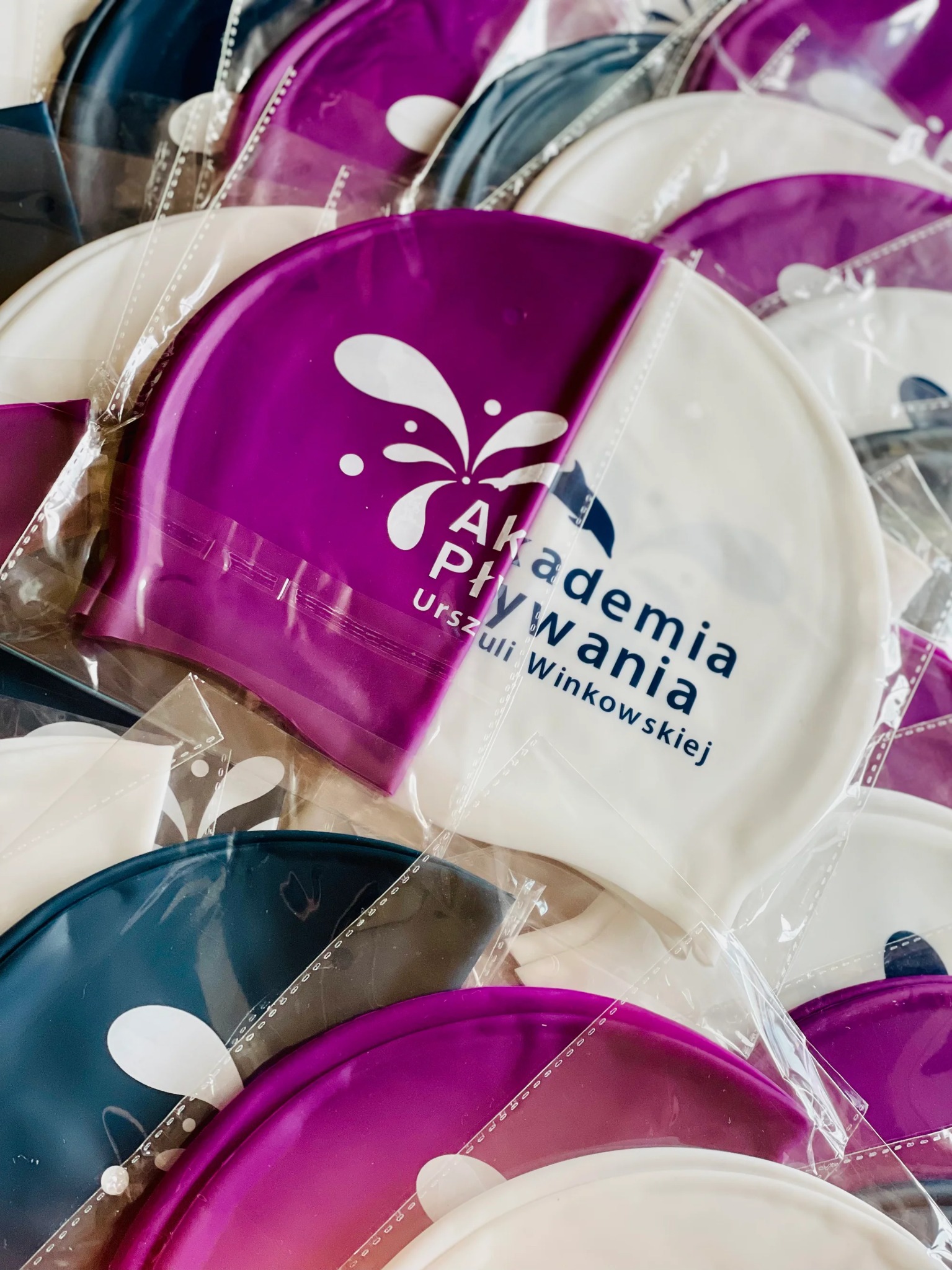 Stos czapeczek pływackich w foliowych opakowaniach, w kolorach białym, fioletowym i granatowym, z nadrukowanym logo i nazwą szkoły pływania 'Akademia Pływania Urszuli Winkowskiej'.