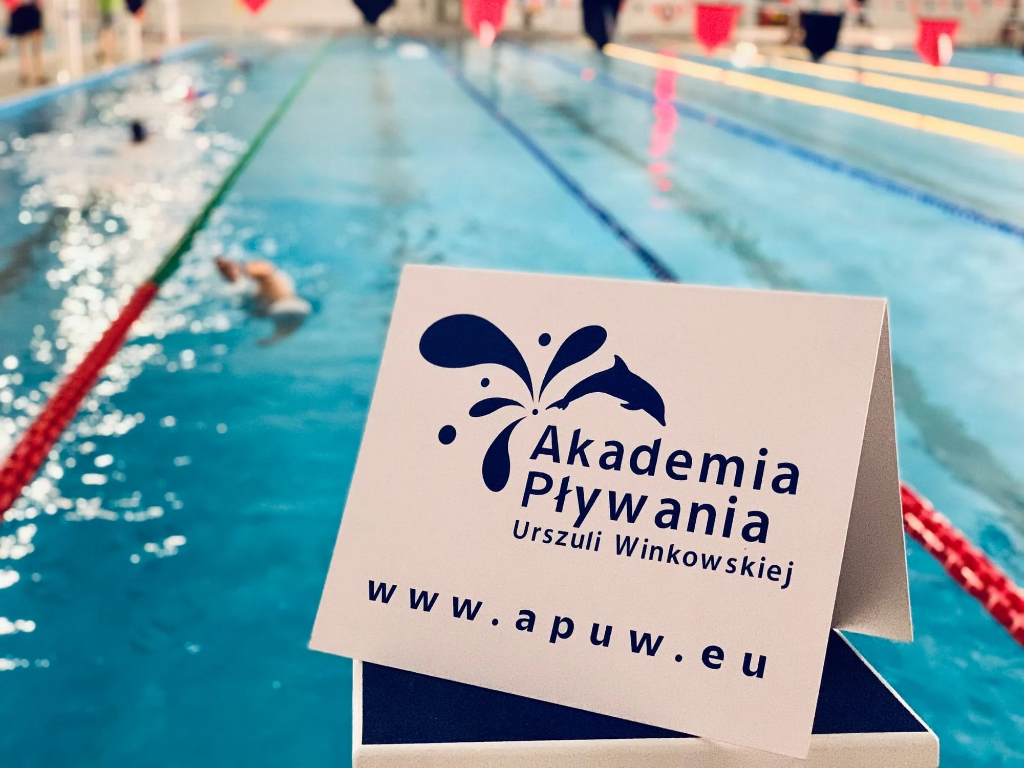 Wizytówka Akademii Pływania Urszuli Winkowskiej na tle basenu z pływającym mężczyzną, widoczne tory pływackie i oznaczenia.