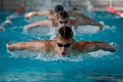Pływak w okularach wykonuje motylka w basenie, za nim dwóch innych pływaków. Widoczne fale i dynamika ruchu.