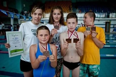 Grupa młodych pływaków z brązowymi medalami i trenerką trzymającą dyplom na tle basenu.