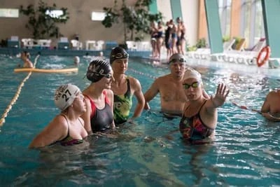 Instruktor pływania w okularach i czepku tłumaczy grupie kursantów w basenie, unosząc dłoń w geście objaśniającym.