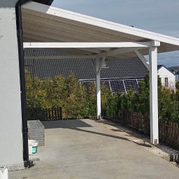 Usługi ogólne budowlanego - Zadaszone miejsce parkingowe z białymi słupami i dachem, widok na sąsiednie domy z panelami słonecznymi i placem zabaw, stolik z naczyniami w lewym dolnym rogu.