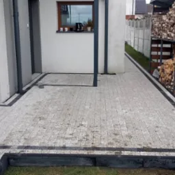 Jasnoszara kostka brukowa z ciemnoszarą obwódką, ułożona na podwórku domu jednorodzinnego, widoczna skrzynka gazowa na drewnianej ścianie i stos drewna oparty o betonowy płot.