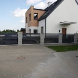 Nowoczesne ogrodzenie panelowe w kolorze grafitowym z betonowymi słupkami, przylegające do dwukondygnacyjnego domu z białymi i beżowymi elewacjami, widoczne z poziomu brukowanego podjazdu.