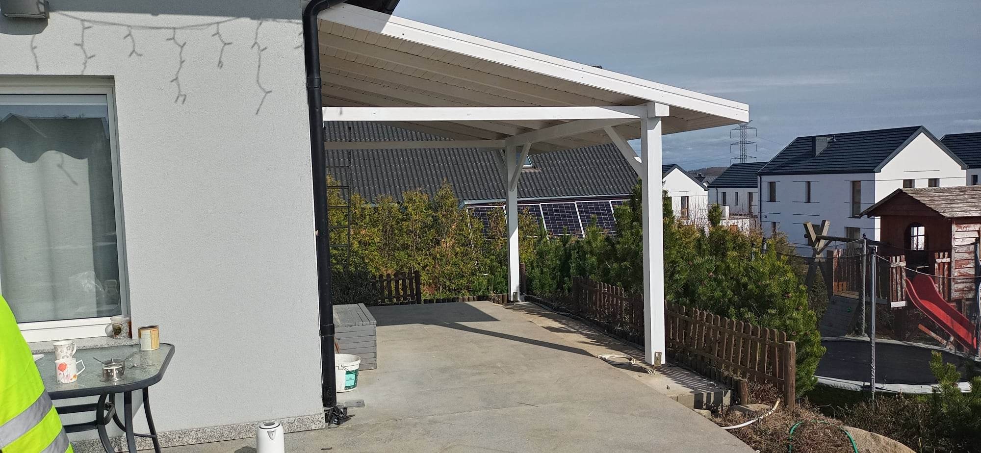 Zadaszone miejsce parkingowe z białymi słupami i dachem, widok na sąsiednie domy z panelami słonecznymi i placem zabaw, stolik z naczyniami w lewym dolnym rogu.