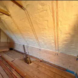 Poddasze w trakcie ocieplania pianką: widoczne drewniane elementy konstrukcyjne, fragment komina z szarego pustaka oraz świeżo nałożona warstwa izolacji z pianki o nierównej fakturze, częściowo...