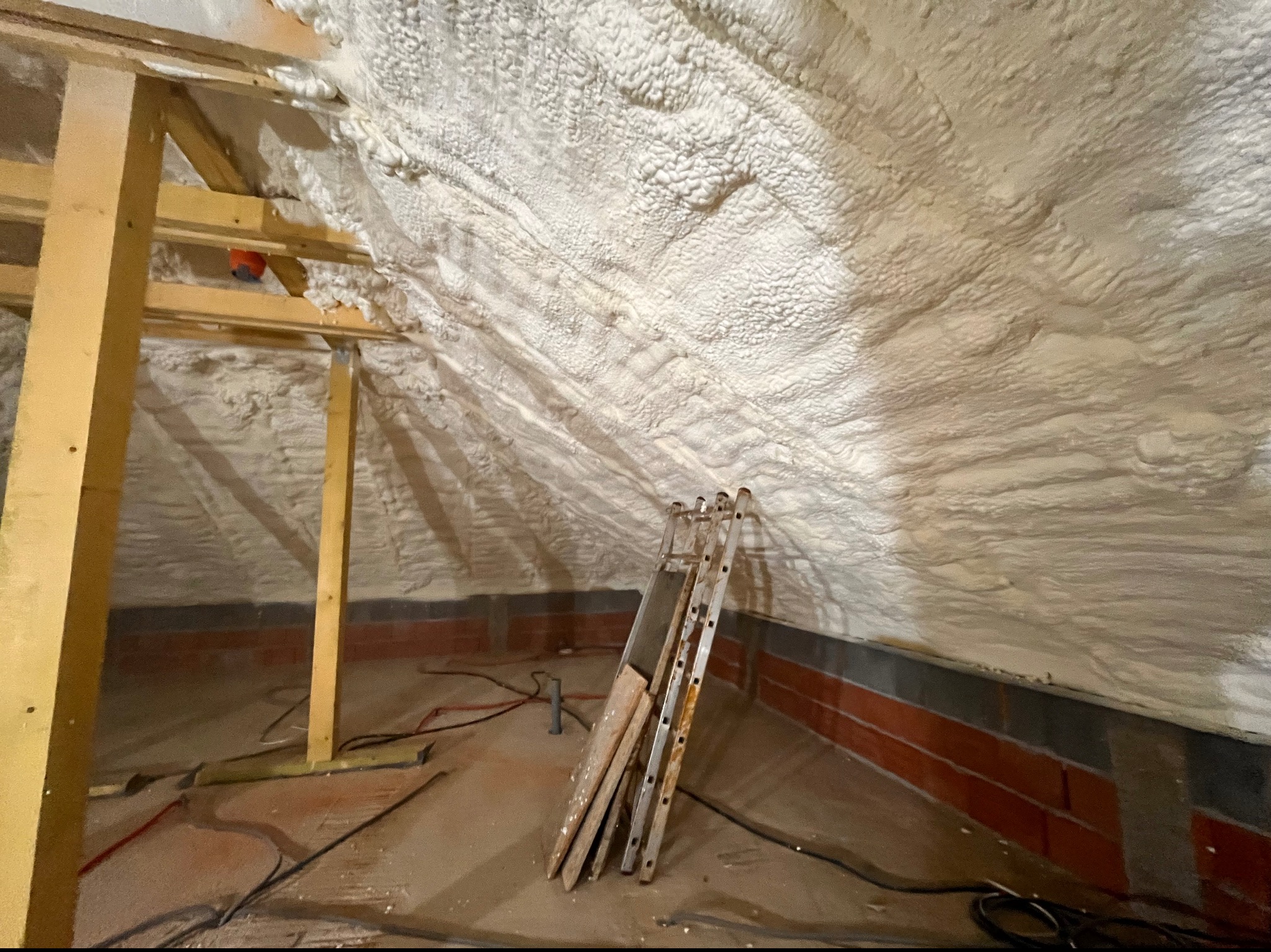 Poddasze z drewnianą konstrukcją i ścianami pokrytymi grubą warstwą białej pianki izolacyjnej, obok ściany oparta drabina, widoczne kable na podłodze.