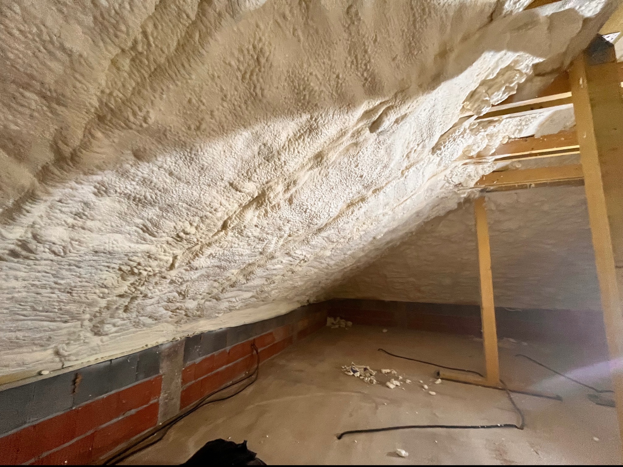 Poddasze po natrysku pianki izolacyjnej, widoczne nierówności i struktura piany, fragment muru z cegły i drewniane elementy konstrukcyjne.